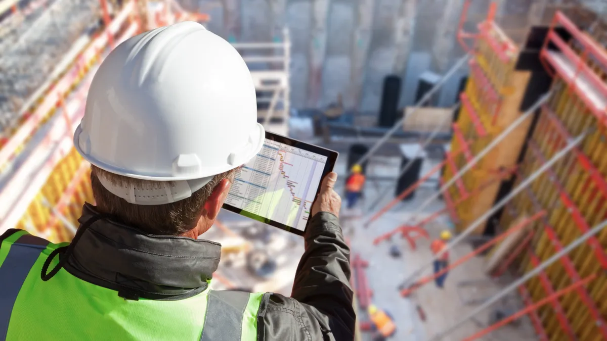 A worker looks down at his tablet while on a construction site. He wears safety gear.