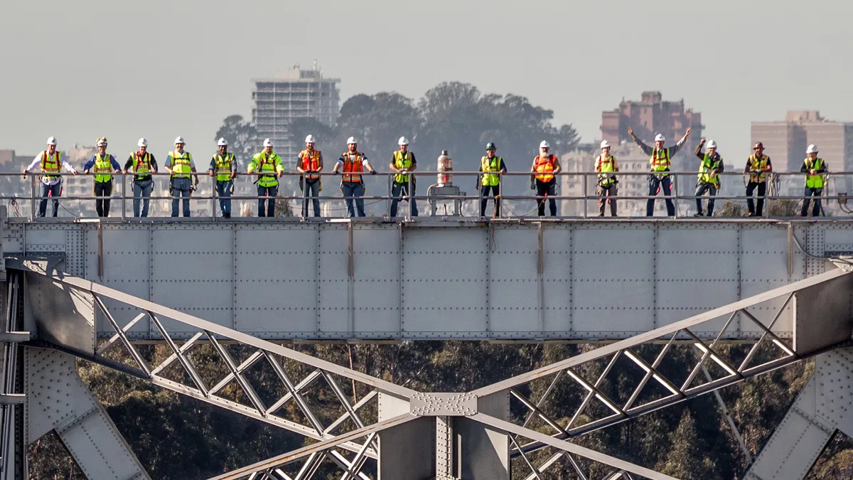 A group of people in safety gear stand atop a metal structure and wave at the camera in the sunset light.