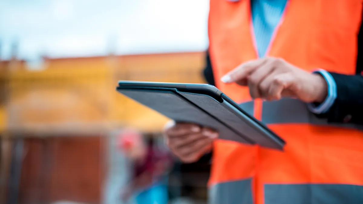 A man in a suit and an orange safety vest uses a tablet on a construction site. The user is in the foreground and clear, the background is blurred.
